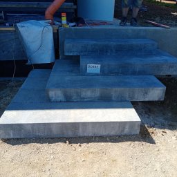 Kiernia AB - Stopnie wejściowe z szarych płyt betonowych w trakcie montażu, widoczne narzędzia i materiały budowlane, w tle fragment elewacji i ogrodzenia.