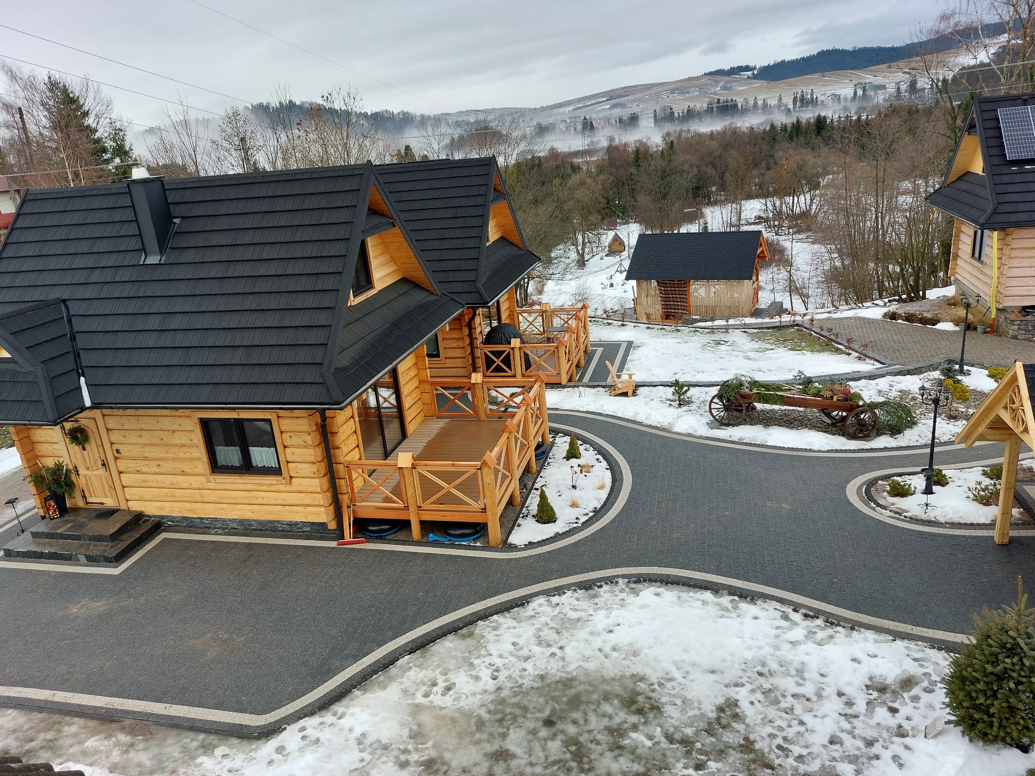 Widok z góry na posesję z drewnianymi domkami w górskim stylu, z czarną kostką brukową tworzącą ścieżki i podjazdy, otoczoną śnieżnym krajobrazem i zalesionymi wzgórzami w tle.