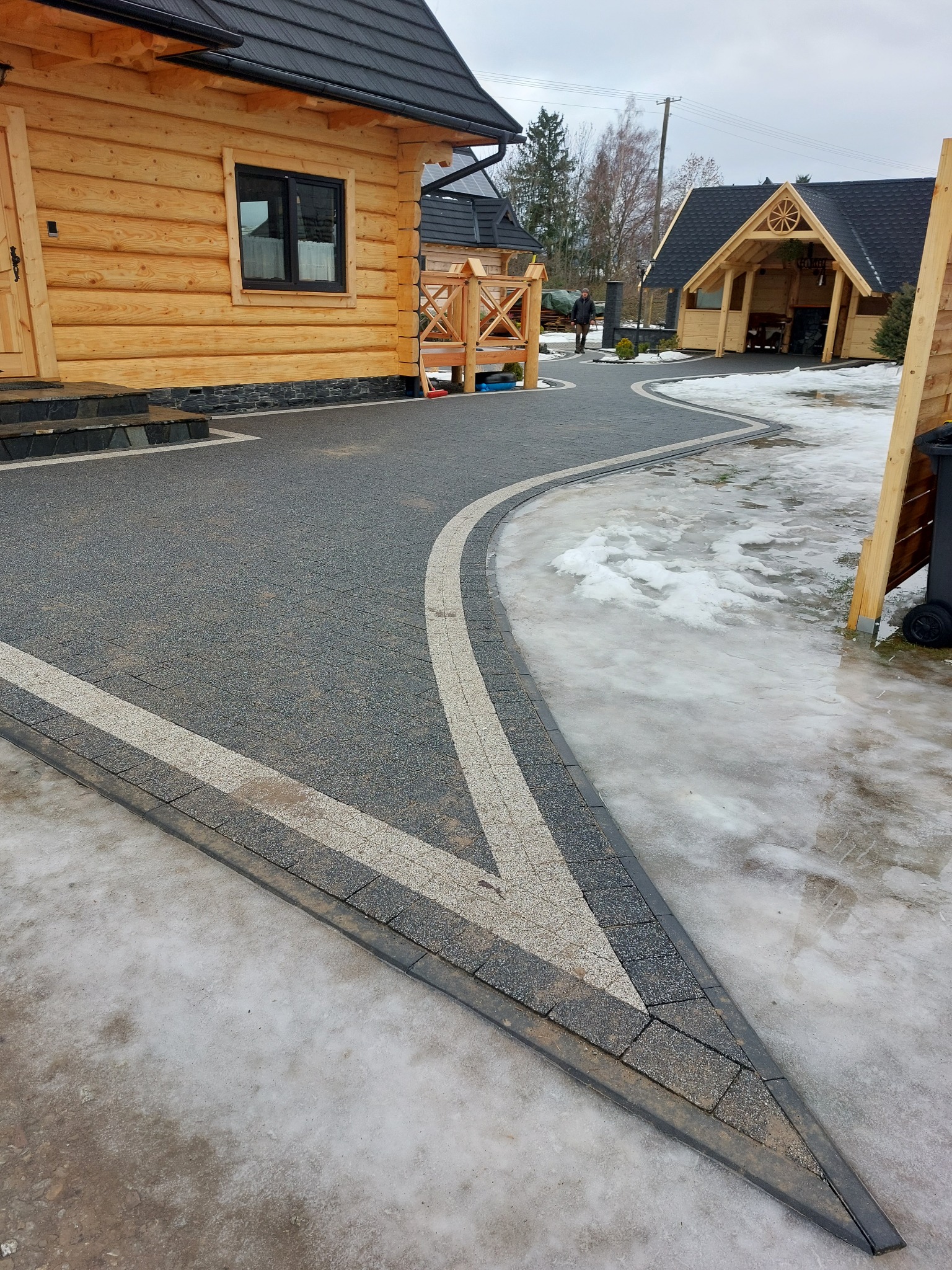 Ułożona kostka brukowa w kolorze szarym z jasnymi pasami, tworząca ścieżkę przed drewnianym domem i altaną, z resztkami śniegu na obrzeżach.