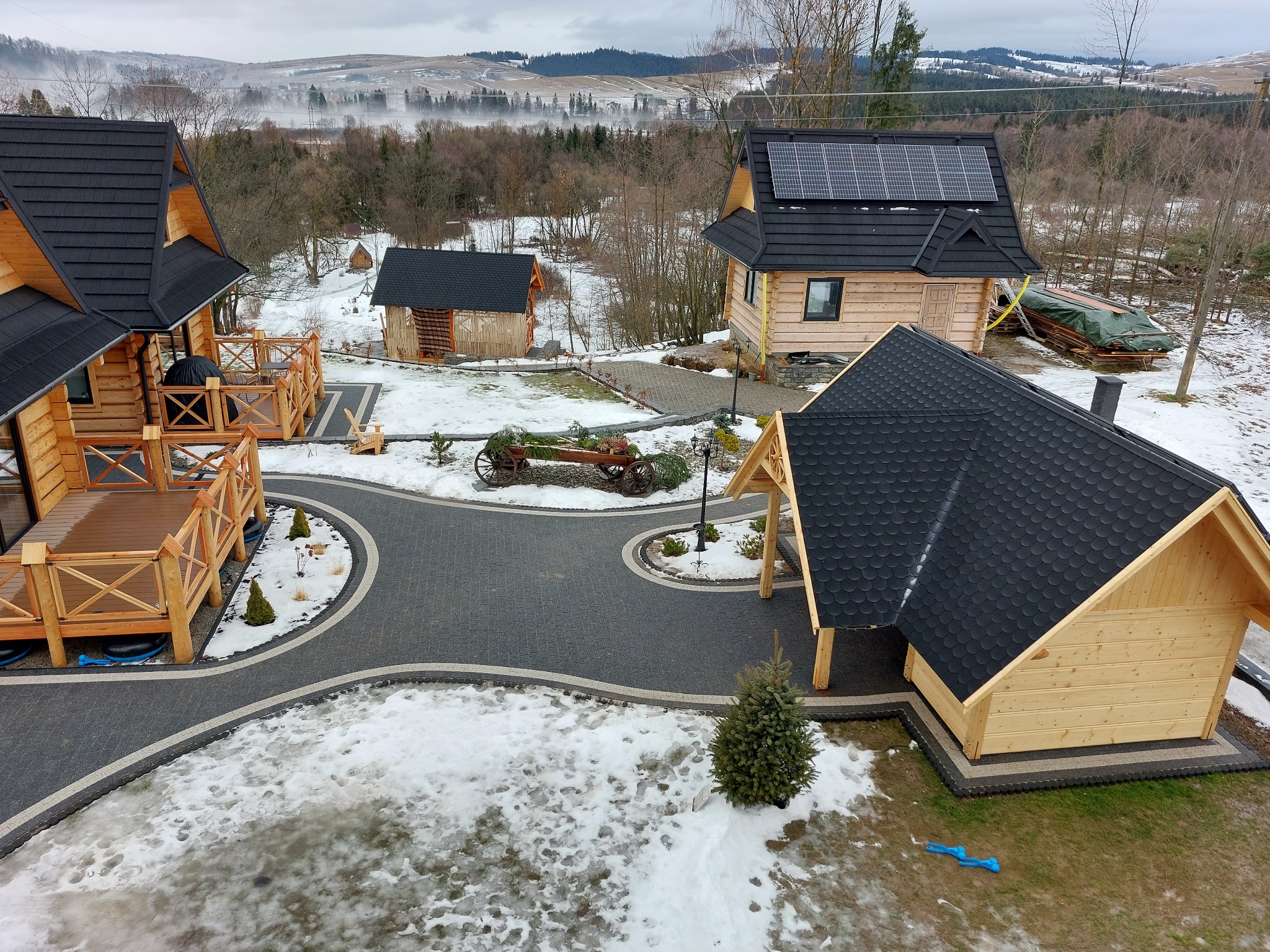 Widok z góry na kompleks drewnianych domków z czarnymi dachami, połączonych ścieżkami z ciemnej kostki brukowej z jasnymi obrzeżami, otoczonych śniegiem i krajobrazem górskim w tle.