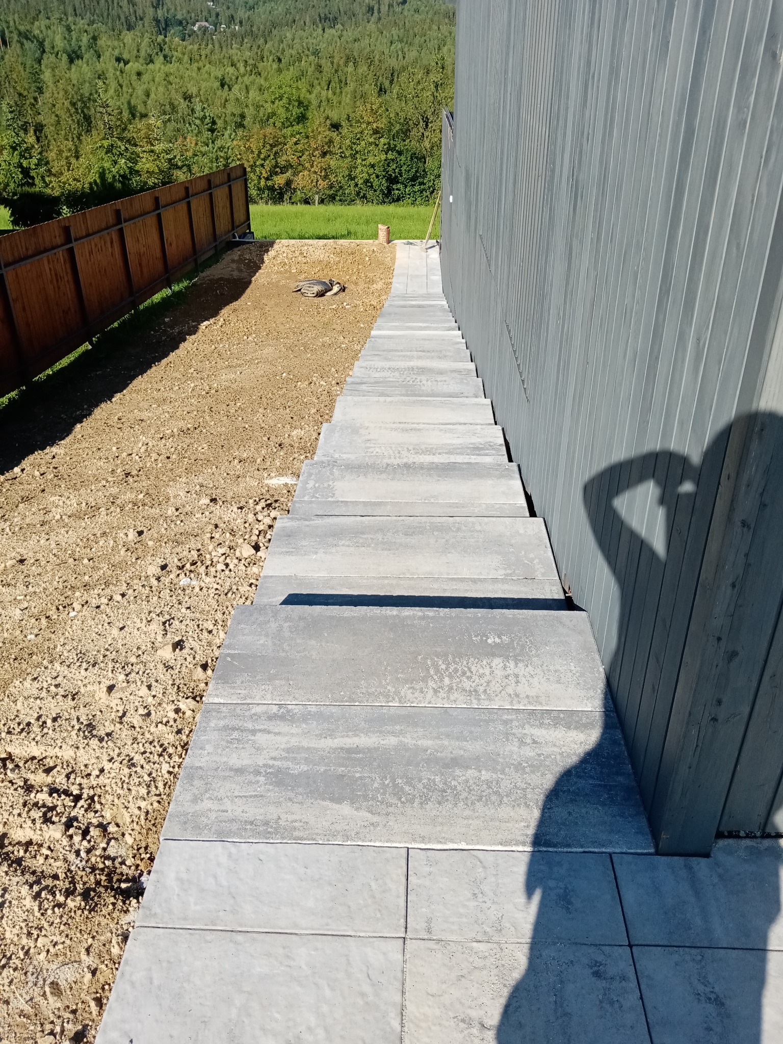 Świeżo ułożona, szara kostka brukowa prowadząca wzdłuż drewnianej ściany budynku, z widocznym przygotowanym terenem pod dalsze prace i cieniem osoby robiącej zdjęcie.