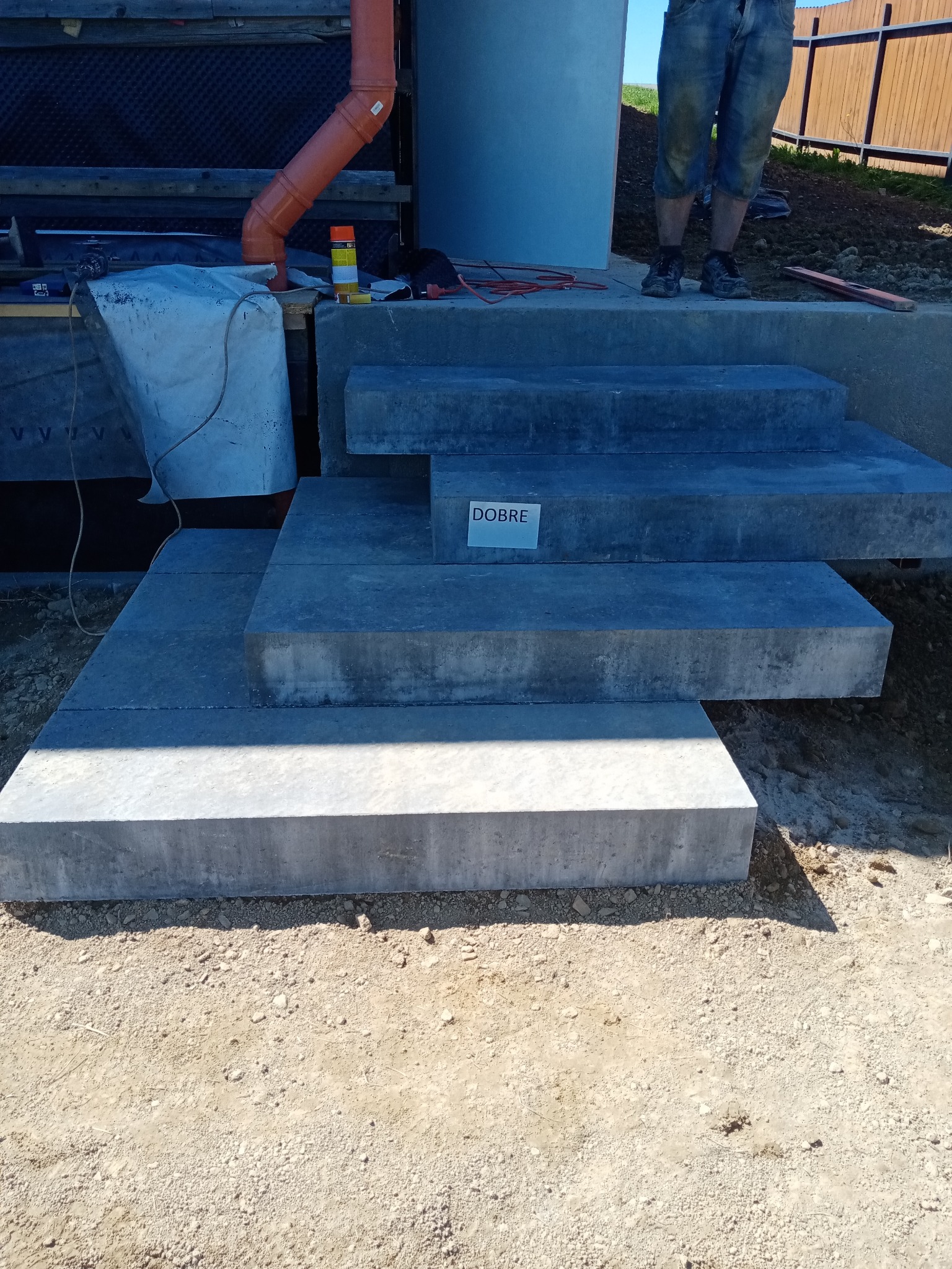 Stopnie wejściowe z szarych płyt betonowych w trakcie montażu, widoczne narzędzia i materiały budowlane, w tle fragment elewacji i ogrodzenia.