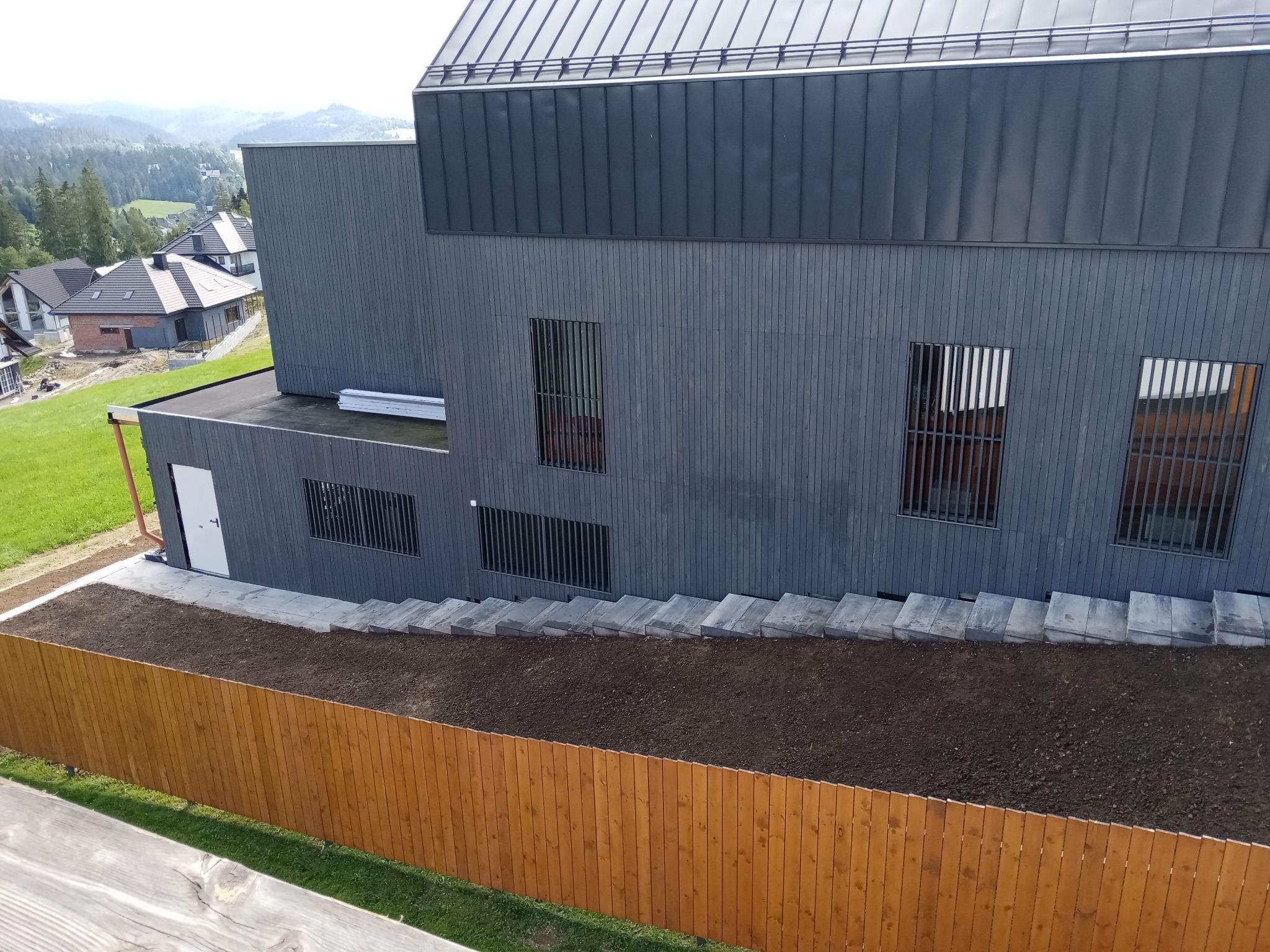 Nowoczesny dom z szarą elewacją, widoczne schody z kostki prowadzące do wejścia, świeżo nawieziona ziemia i drewniany płot w tle, krajobraz górski w oddali.