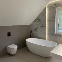 Nowoczesna łazienka na poddaszu z wolnostojącą białą wanną, umywalką nablatową i podwieszaną toaletą, wykończona płytkami imitującymi marmur z oświetleniem LED.