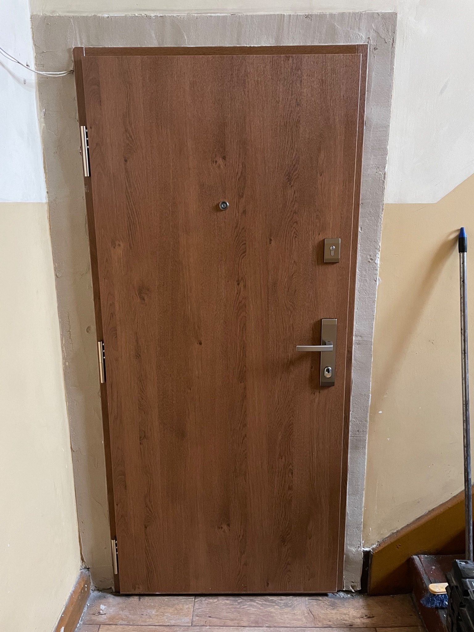 Brązowe drzwi wejściowe z srebrną klamką i wizjerem, osadzone w ościeżnicy z surowym tynkiem, widoczne ślady po montażu, podłoga z drewnopodobnych paneli.