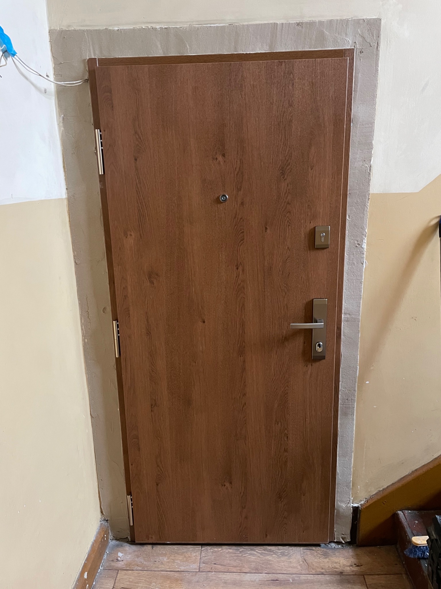 Zamknięte, brązowe drzwi wejściowe z wizjerem i srebrną klamką, osadzone w ościeżnicy z widocznymi śladami świeżego tynku, ukazujące etap montażu w surowym wnętrzu klatki schodowej.