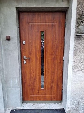 Brązowe drzwi wejściowe z pionowym przeszkleniem, widoczne odbicie osoby w szybie, szara ościeżnica i fragment ściany z dzwonkiem.