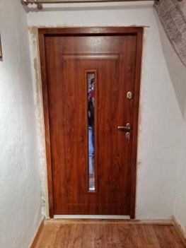 Brązowe drzwi wejściowe z podłużnym przeszkleniem, widoczne ślady po montażu na ościeżnicy, jasne ściany i podłoga.