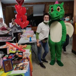 Animator w kostiumie zielonego smoka pozuje z kobietą w maseczce na tle stołu z rekwizytami do malowania twarzy i balonami w kształcie serc.