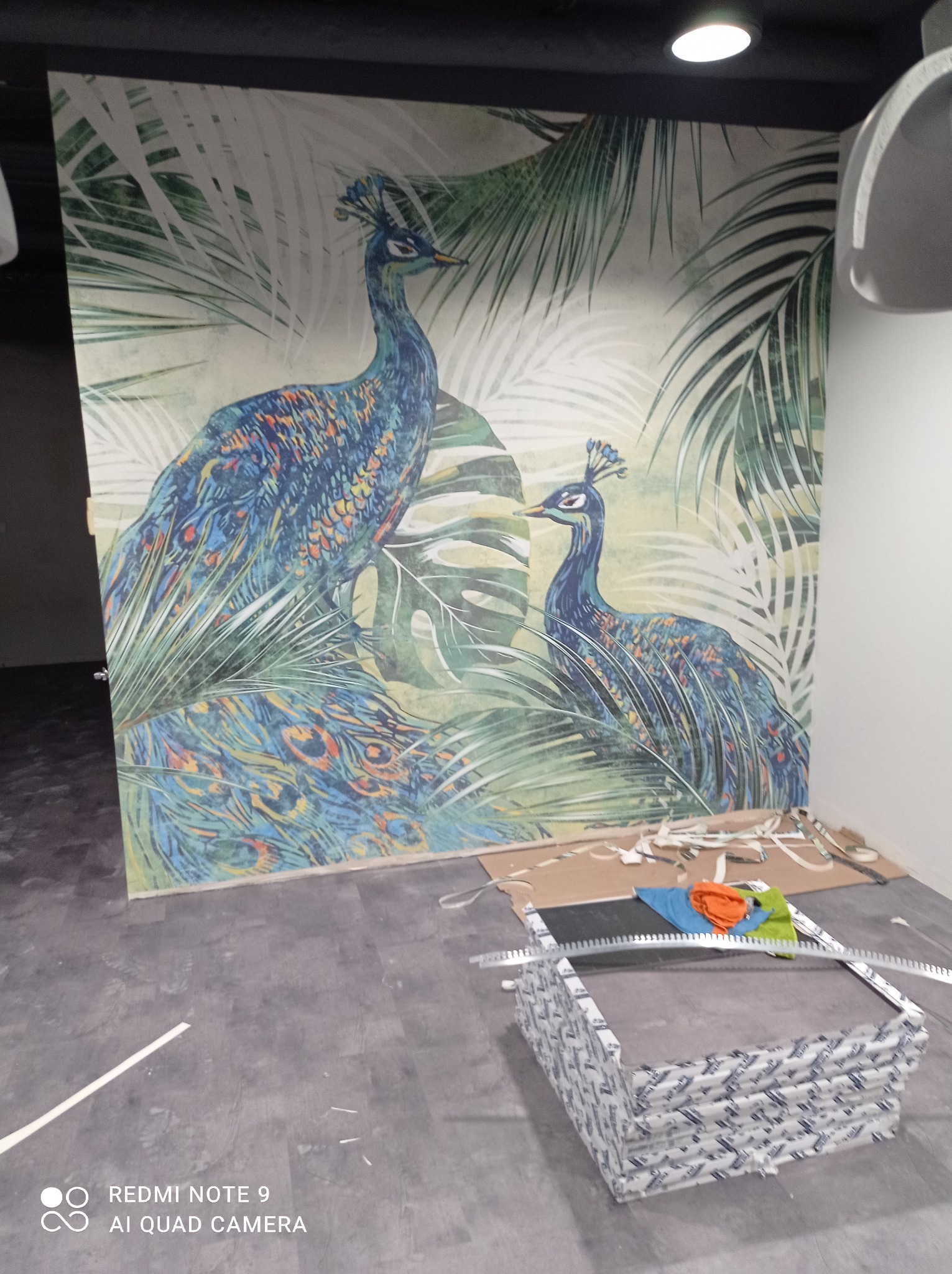 Świeżo położona tapeta z motywem pawia i tropikalnych liści w pomieszczeniu, widoczne narzędzia i pozostałości po pracy.