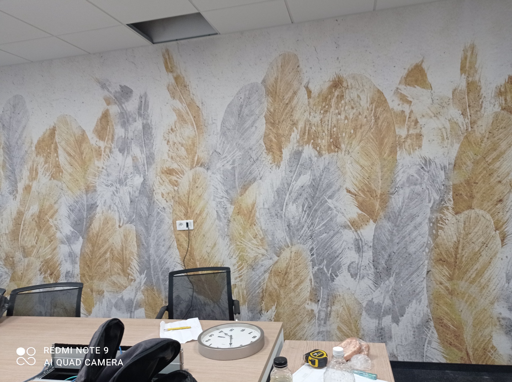 Wzorzysta tapeta z motywem piór w odcieniach złota i szarości na ścianie biura, widoczne biurko, krzesła i akcesoria biurowe