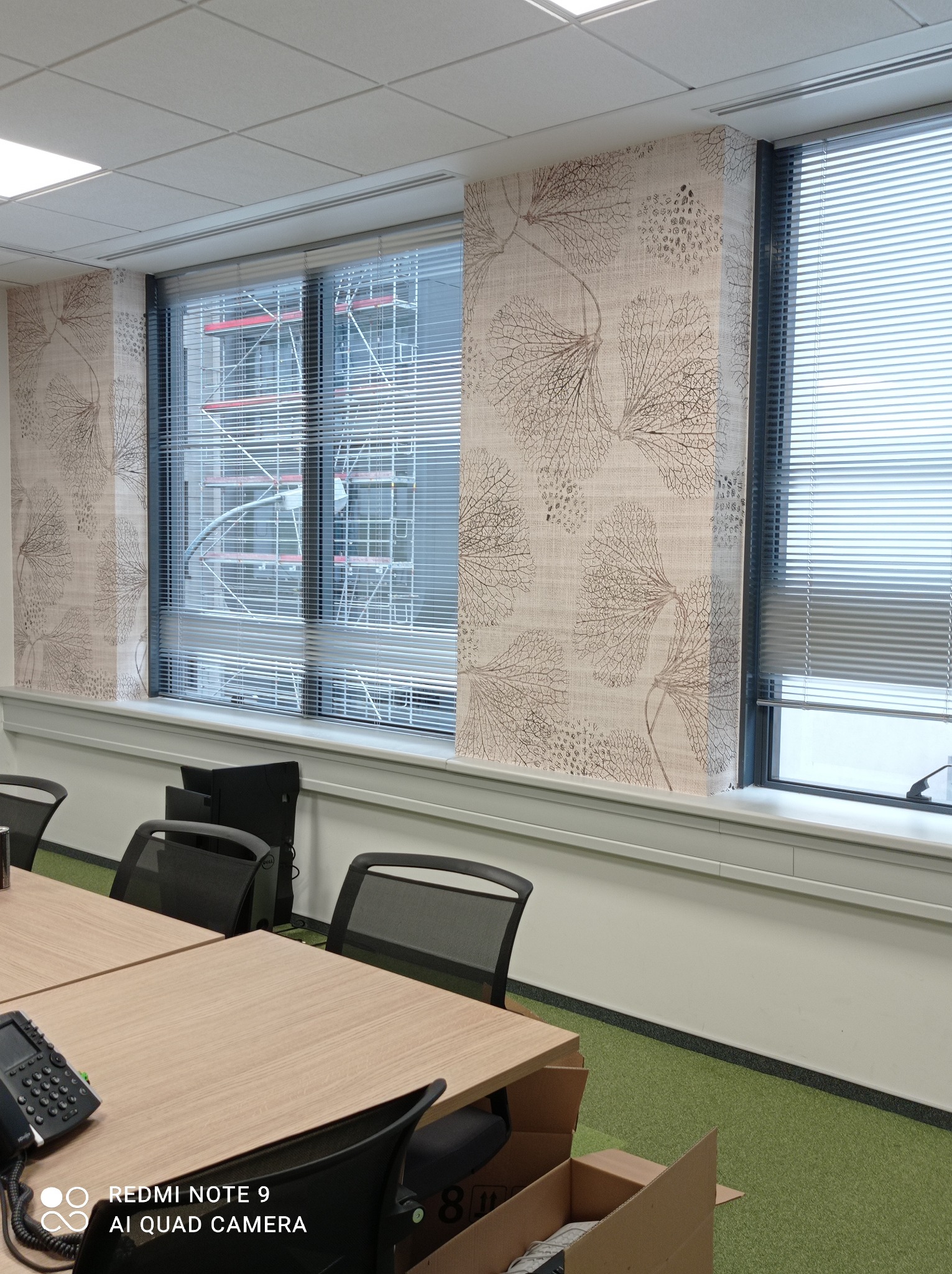 Biuro po remoncie: ściany z tapetą w delikatny wzór liści, okna z żaluzjami, widok na rusztowania za oknem, biurko konferencyjne z krzesłami.