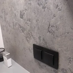 Ściana z szarą, dekoracyjną fakturą imitującą kamień, z zamontowanym czarnym, podwójnym przyciskiem spłuczki; obok widoczny flakon perfum i kosz na śmieci.