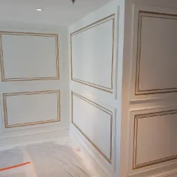 Białe ściany z dekoracyjnymi, prostokątnymi panelami z delikatnym złotym obramowaniem, zabezpieczona podłoga folią malarską.