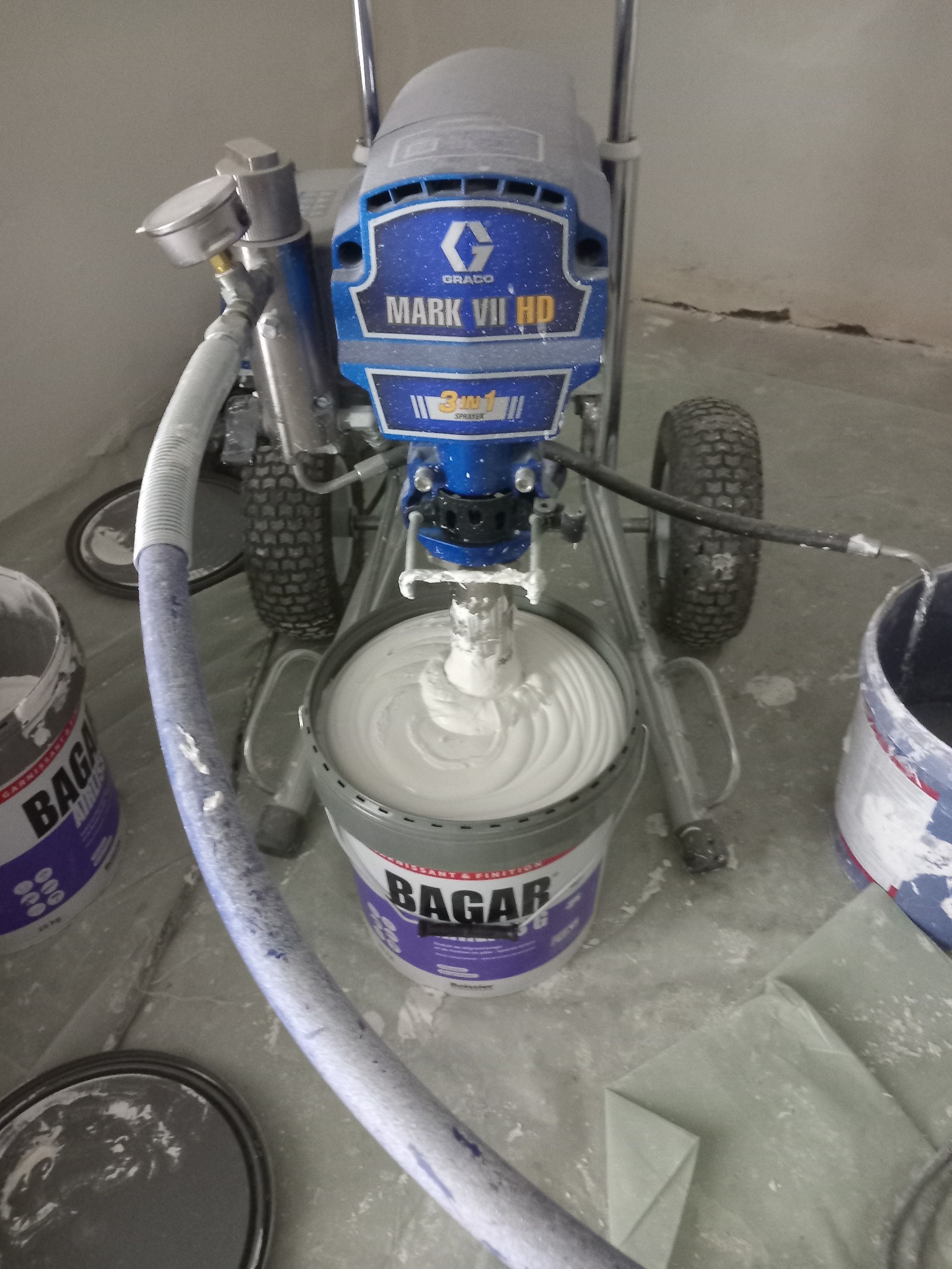 Urządzenie Graco Mark VII HD zanurzone w wiadrze z gładzią szpachlową Bagar, otoczone wiadrami i folią ochronną na podłodze, sugerujące przygotowanie do prac wykończeniowych.