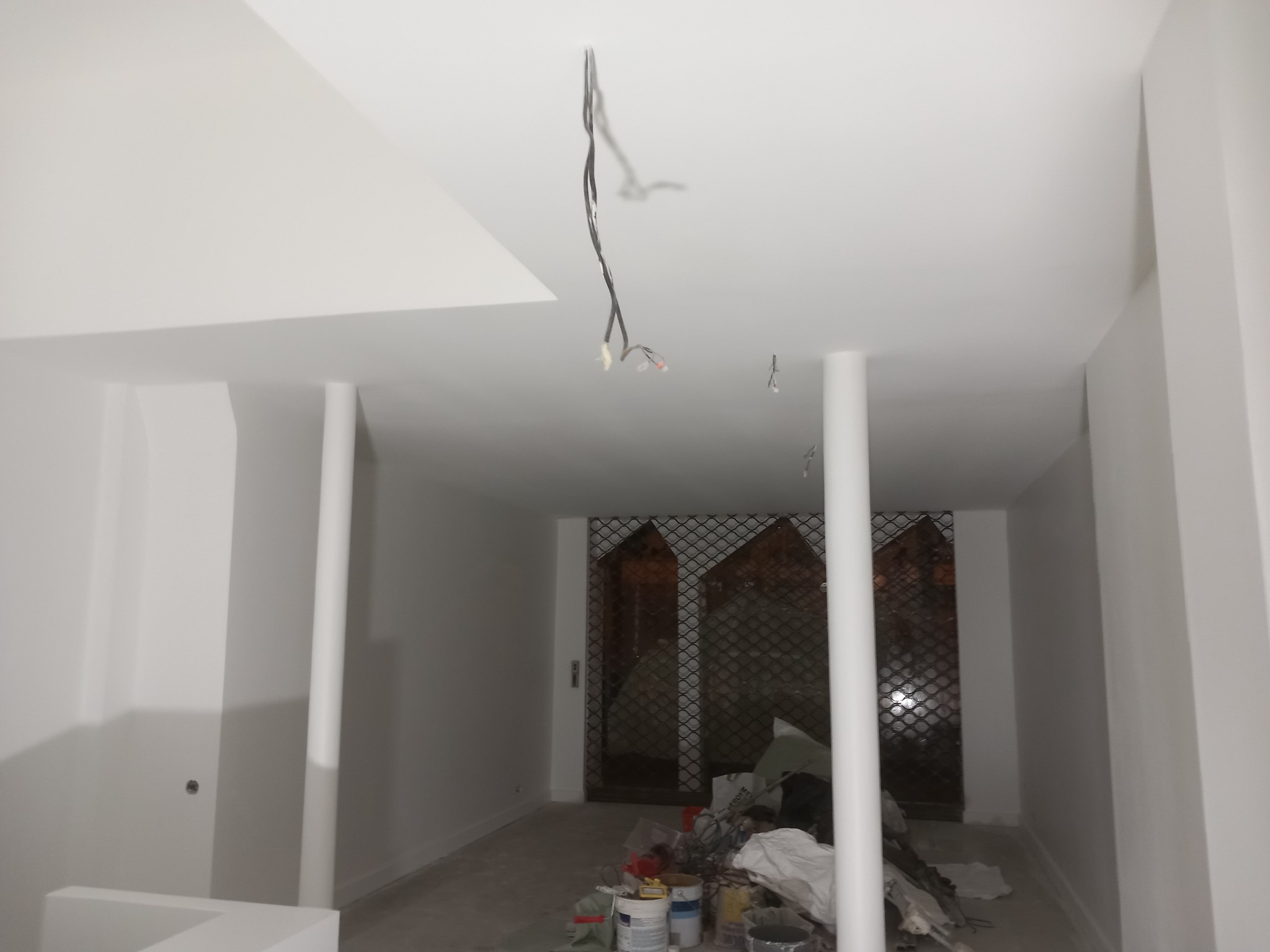Wnętrze w trakcie prac wykończeniowych: świeżo pomalowane białe ściany i sufit, widoczne kable zwisające z sufitu, kolumny, a w tle zabezpieczone kratą drzwi i bałagan po pracach malarskich.