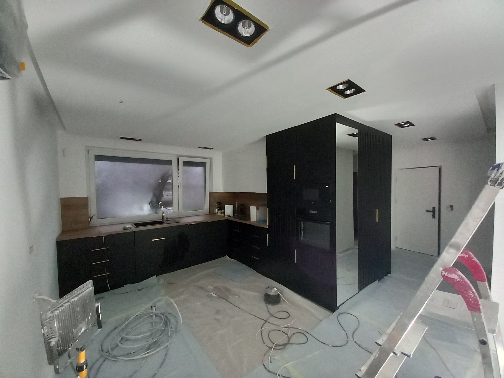 Wnętrze kuchni w trakcie prac wykończeniowych: czarne szafki, drewniany blat, podłoga zabezpieczona folią, widoczny grzejnik i drabina, surowe ściany przygotowane do malowania.