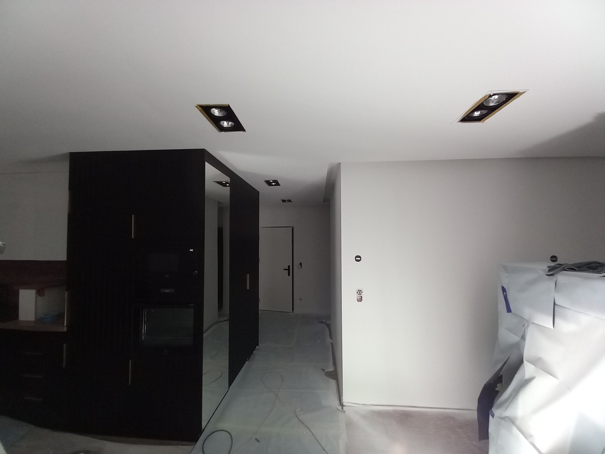 Wnętrze mieszkania w trakcie prac wykończeniowych: świeżo pomalowane ściany, zabezpieczona podłoga folią ochronną, widoczne puszki elektryczne i oprawy oświetleniowe w suficie.