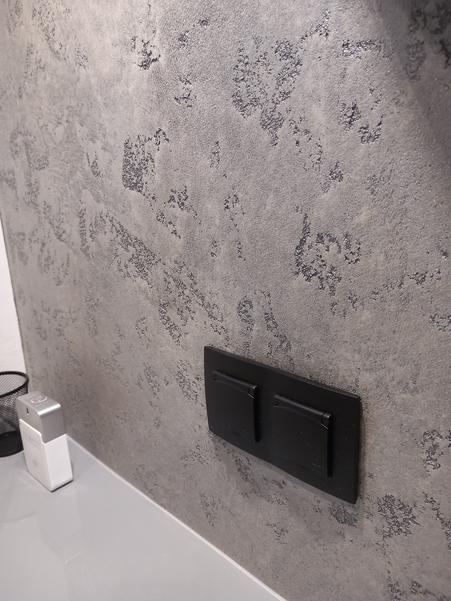 Ściana z szarą, dekoracyjną fakturą imitującą kamień, z zamontowanym czarnym, podwójnym przyciskiem spłuczki; obok widoczny flakon perfum i kosz na śmieci.