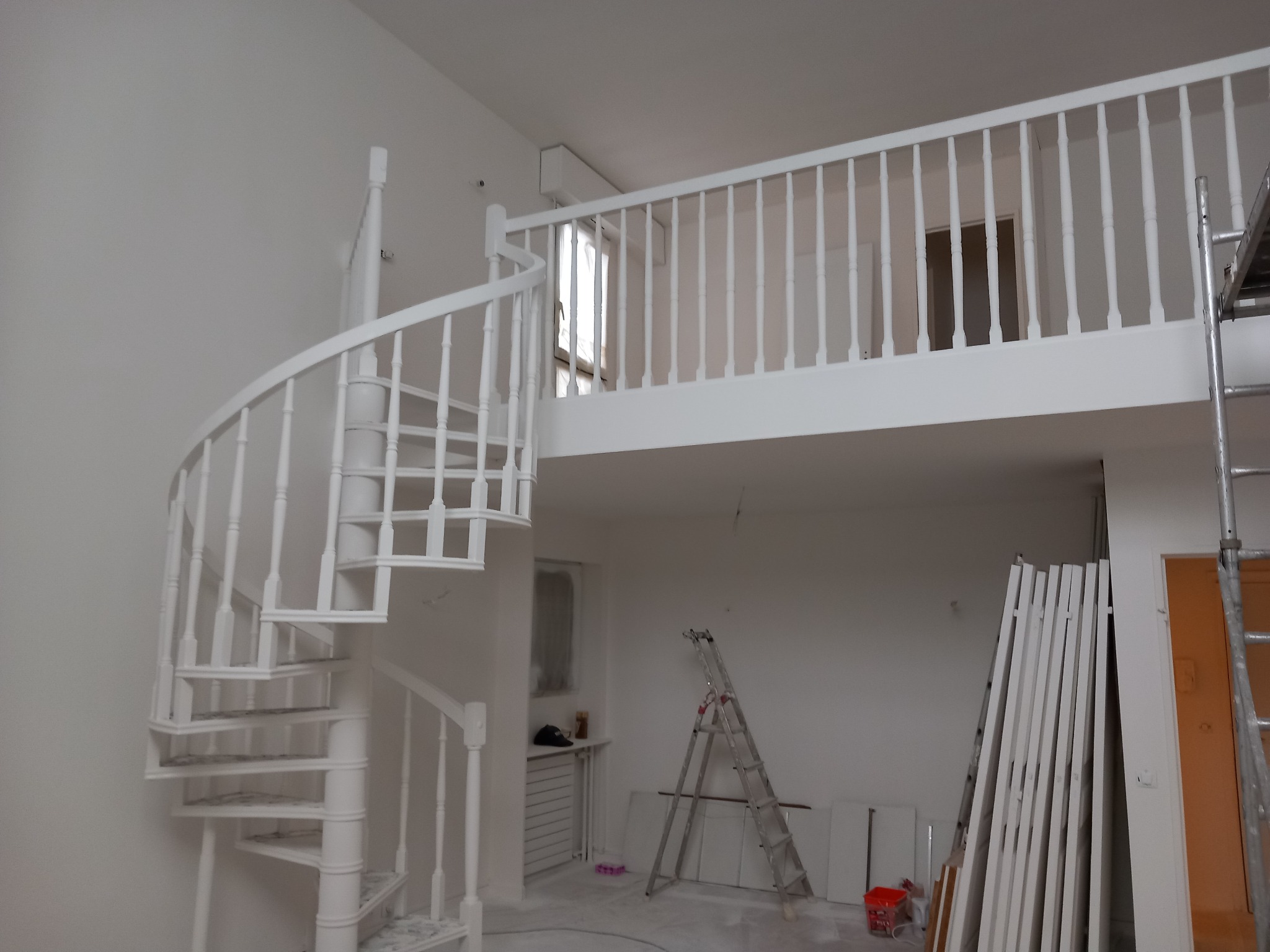 Wnętrze w trakcie prac wykończeniowych: białe spiralne schody, balustrada na antresoli i drabina, świeżo pomalowane ściany, elementy do montażu oparte o ścianę.