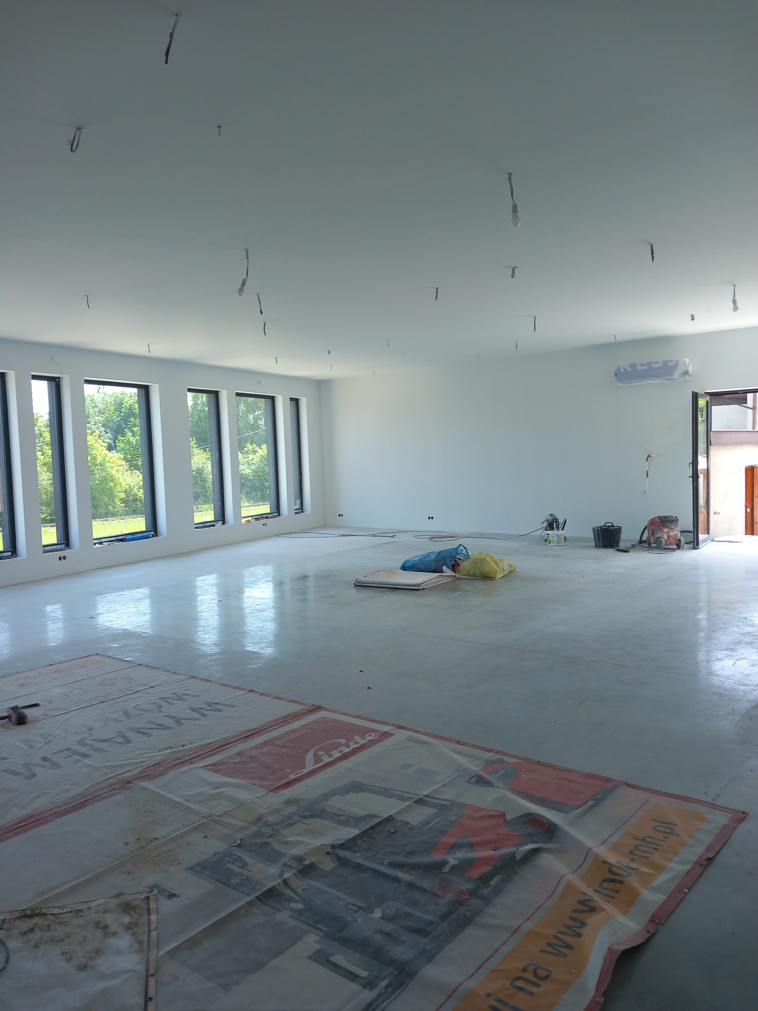 Duże, jasne pomieszczenie w trakcie prac wykończeniowych; widoczne okna, gołe żarówki zwisające z sufitu, zabezpieczona podłoga, worki z materiałami budowlanymi oraz sprzęt do szlifowania i wiadro.