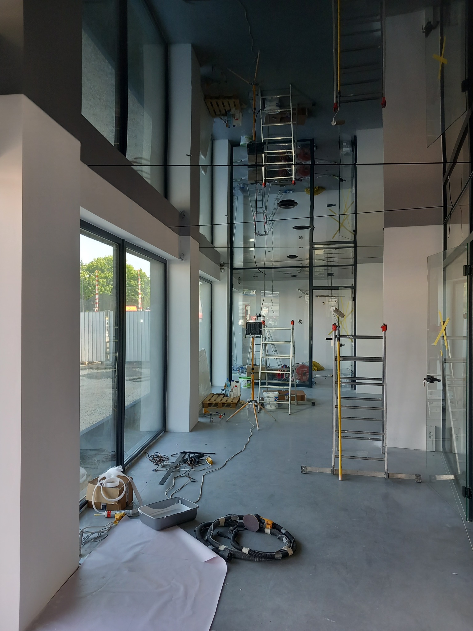 Wnętrze biurowe w trakcie wykańczania: widoczne drabiny, narzędzia malarskie, szpachla, okna z widokiem na zewnątrz, przygotowane do malowania ściany.