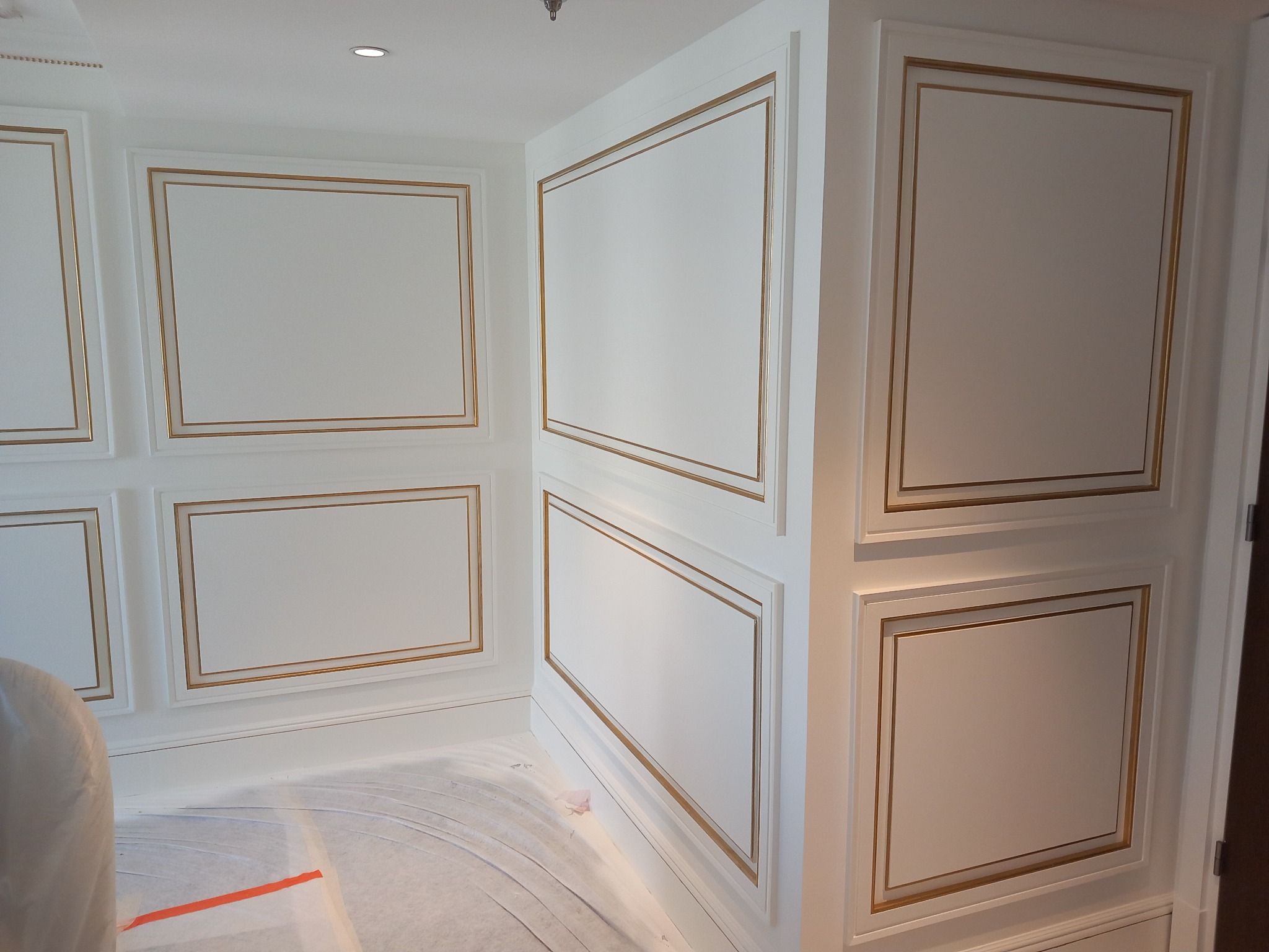 Białe ściany z dekoracyjnymi, prostokątnymi panelami z delikatnym złotym obramowaniem, zabezpieczona podłoga folią malarską.