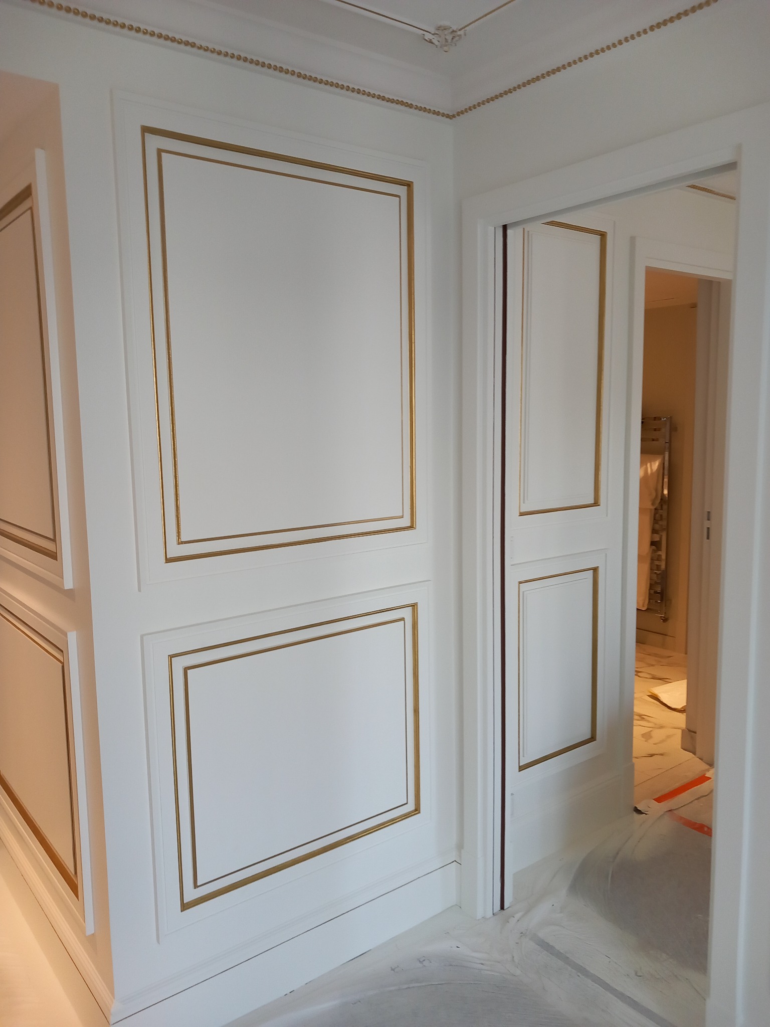 Białe ściany z dekoracyjnymi, złotymi obramowaniami w klasycznym stylu, widoczne zabezpieczenia podłogi folią malarską.