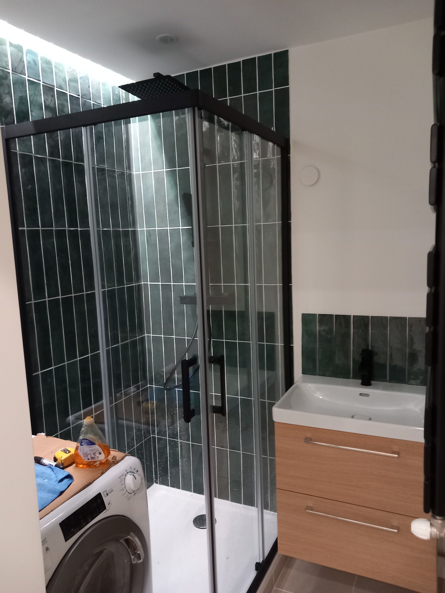 Nowoczesna łazienka po renowacji: kabina prysznicowa z czarnymi profilami i deszczownicą, umywalka z szafką w odcieniach drewna, zielone płytki na ścianach, pralka z narzędziami na górze.