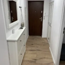 Jasne panele podłogowe w przedpokoju z białymi ścianami, szafką na buty z lustrem, brązowe drzwi wejściowe.