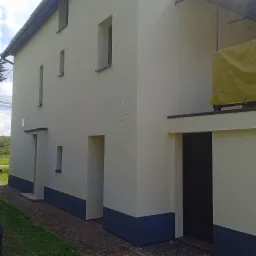 Odnowiona elewacja budynku z kremową farbą, ciemnoszara podmurówka, widoczne okna o nietypowym kształcie i zabezpieczona folią plandeka rusztowania.