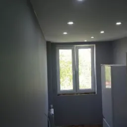 Pomieszczenie pomalowane na szaro z białym oknem, widoczne punktowe oświetlenie sufitowe i fragment białej szafy, grzejnik pod oknem.