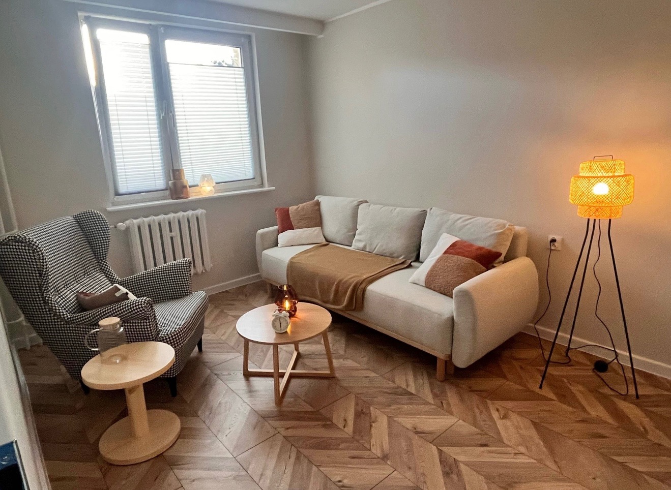 Jasny pokój z sofą, fotelem w pepitkę, stolikiem kawowym z zegarem i lampą stojącą. Podłoga z parkietu w jodełkę. Przytulne wnętrze gabinetu psychologicznego.