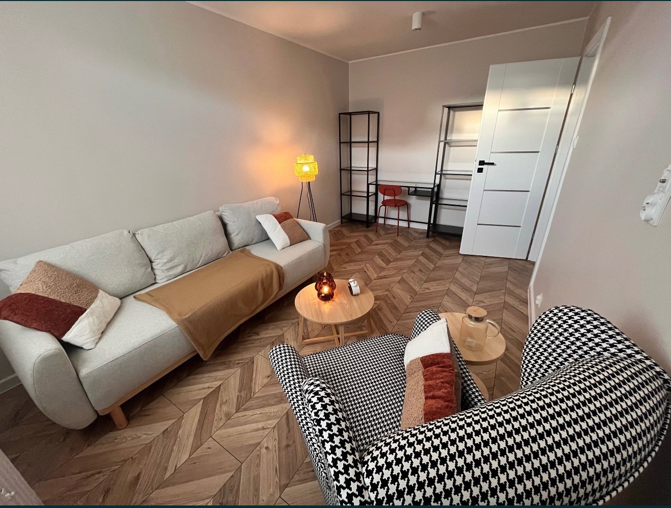 Jasne wnętrze z sofą, wzorzystym fotelem i drewnianą podłogą w jodełkę. Na stoliku świeca i dzbanek. W tle regały i biurko z krzesłem.