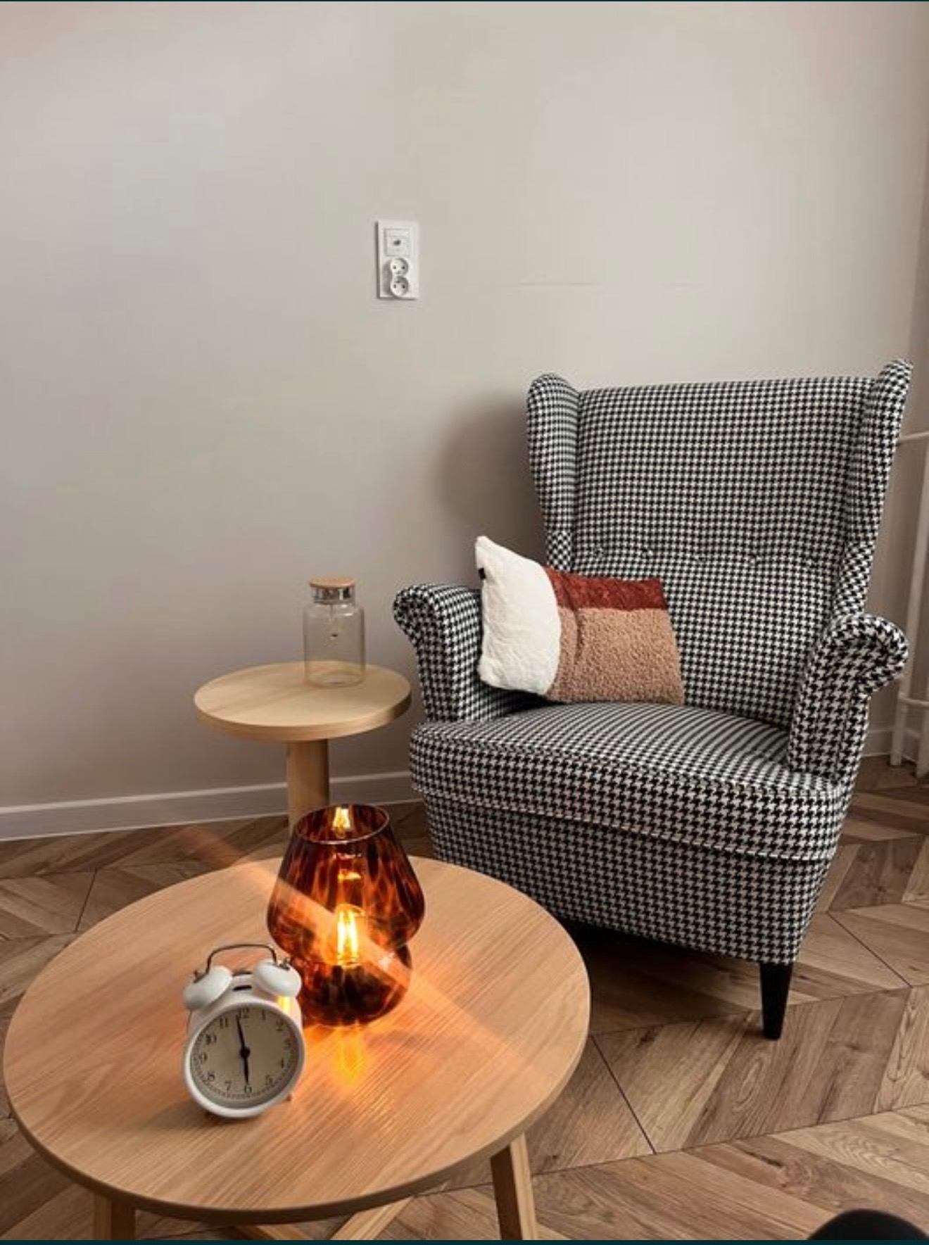 Pokój z fotelem w pepitkę, drewnianymi stolikami i lampą. Na stoliku stoi biały budzik. Fotel z poduszką. Parkiet ułożony w jodełkę. Neutralne kolory.