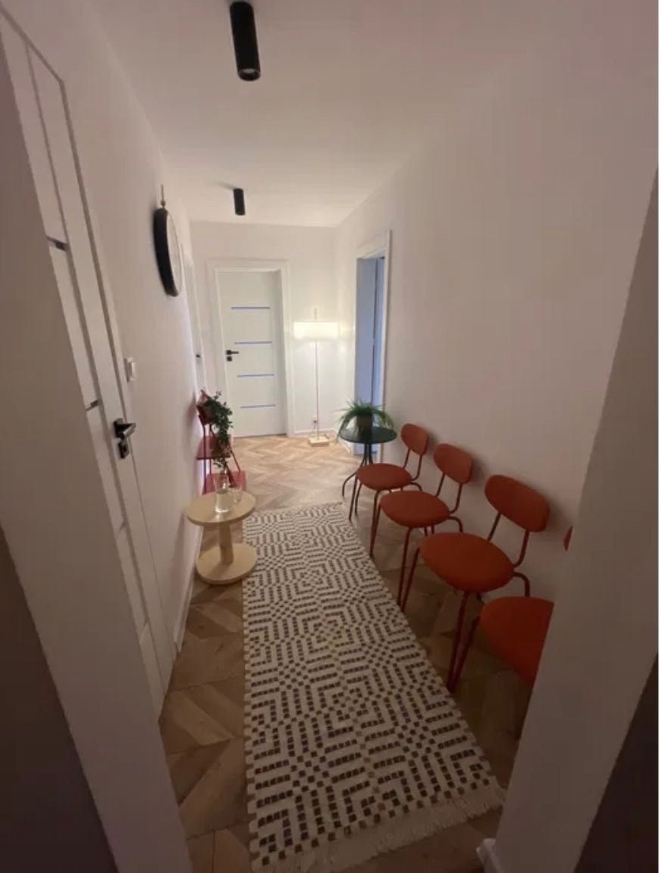 Jasny korytarz z czterema pomarańczowymi krzesłami, dywanem w geometryczne wzory i parkietem w jodełkę. W tle widoczna lampa podłogowa i drzwi.
