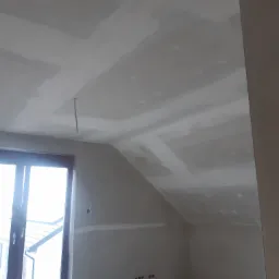 Poddasze ze skośnym sufitem w trakcie prac wykończeniowych, widoczne płyty kartonowo-gipsowe i świeżo nałożone gładzie, surowy przewód zwisający z sufitu, okno dachowe wpuszczające jasne światło.