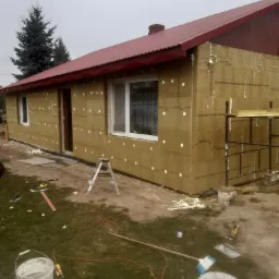 Dom jednorodzinny w trakcie ocieplania ścian zewnętrznych wełną mineralną, widoczne rusztowanie, drabina i narzędzia na trawniku, czerwony dach, pochmurne niebo.