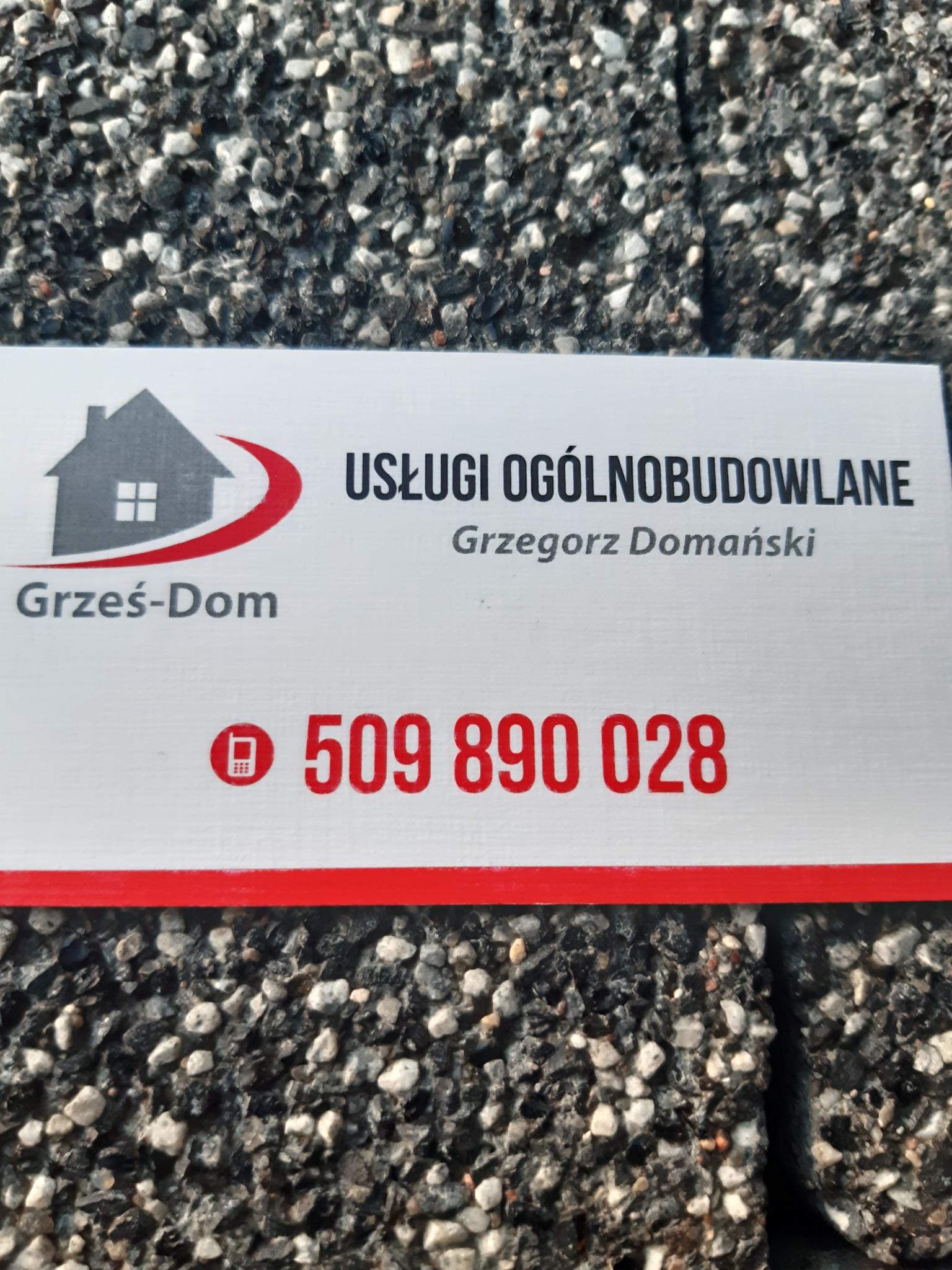 Wizytówka firmy Grześ-Dom oferującej usługi ogólnobudowlane, z numerem telefonu 509 890 028, leżąca na szorstkiej powierzchni.