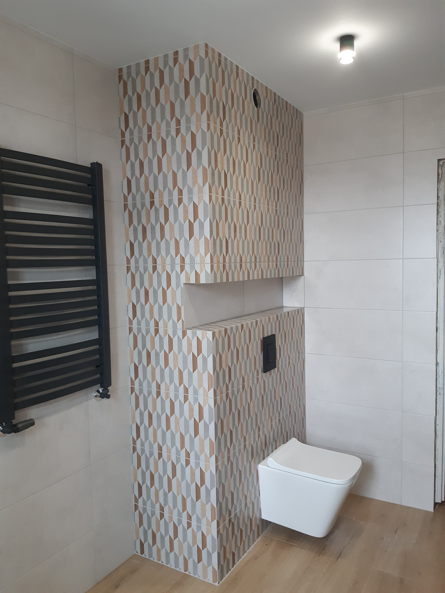 Nowoczesna łazienka z podwieszaną toaletą, grzejnikiem drabinkowym i ścianką z płytek w geometryczny wzór, oświetlona punktowym światłem sufitowym.