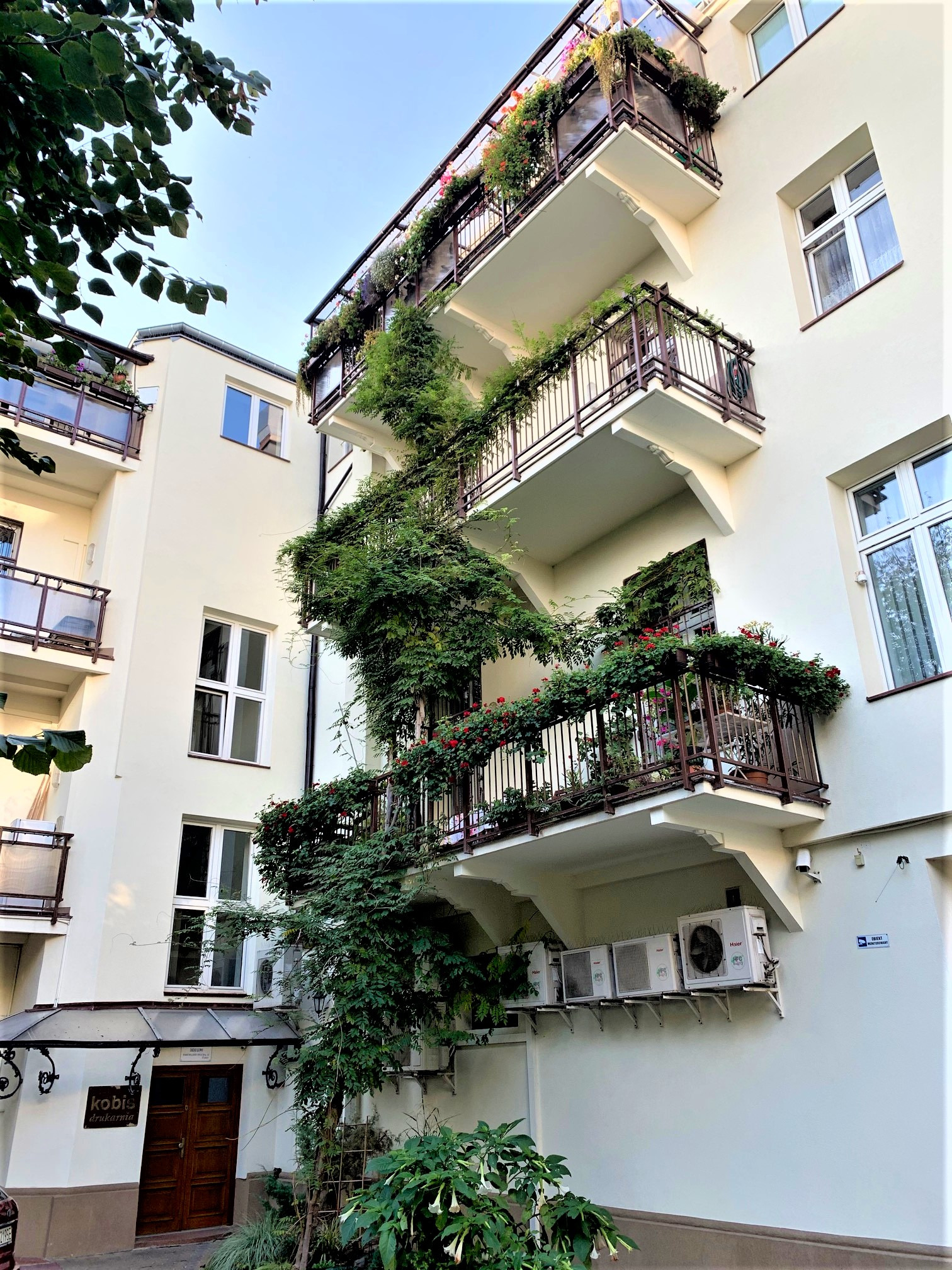 Elewacja budynku apartamentowego z balkonami porośniętymi zielenią i kwiatami, widok z dołu, klimatyzatory na ścianie.