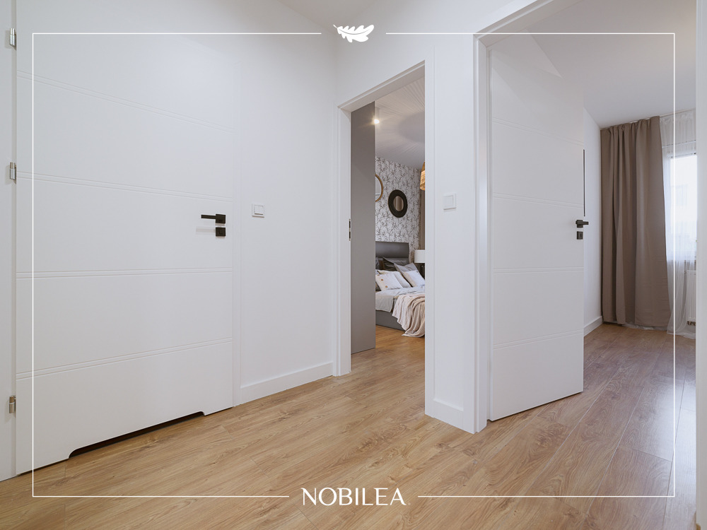 Nowoczesne białe drzwi wewnętrzne z czarnymi klamkami, widok na sypialnię z drewnianą podłogą.