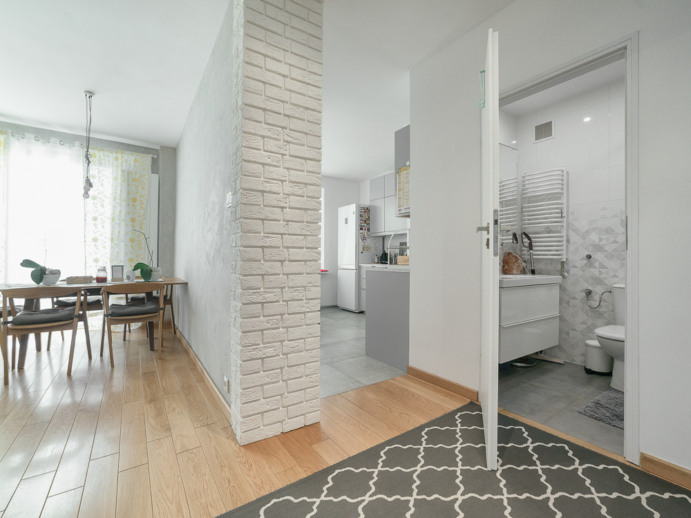 Jasne, przestronne wnętrze mieszkania z drewnianą podłogą, ceglaną ścianą działową, stołem jadalnym i otwartymi drzwiami do łazienki, widok na kuchnię w tle.