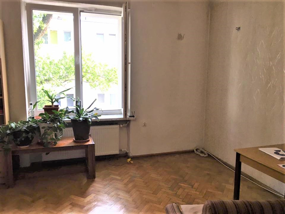 Pusta przestrzeń w mieszkaniu w Warszawie z drewnianą podłogą w jodełkę, stolikiem z roślinami doniczkowymi pod oknem i pustymi ścianami czekającymi na aranżację.