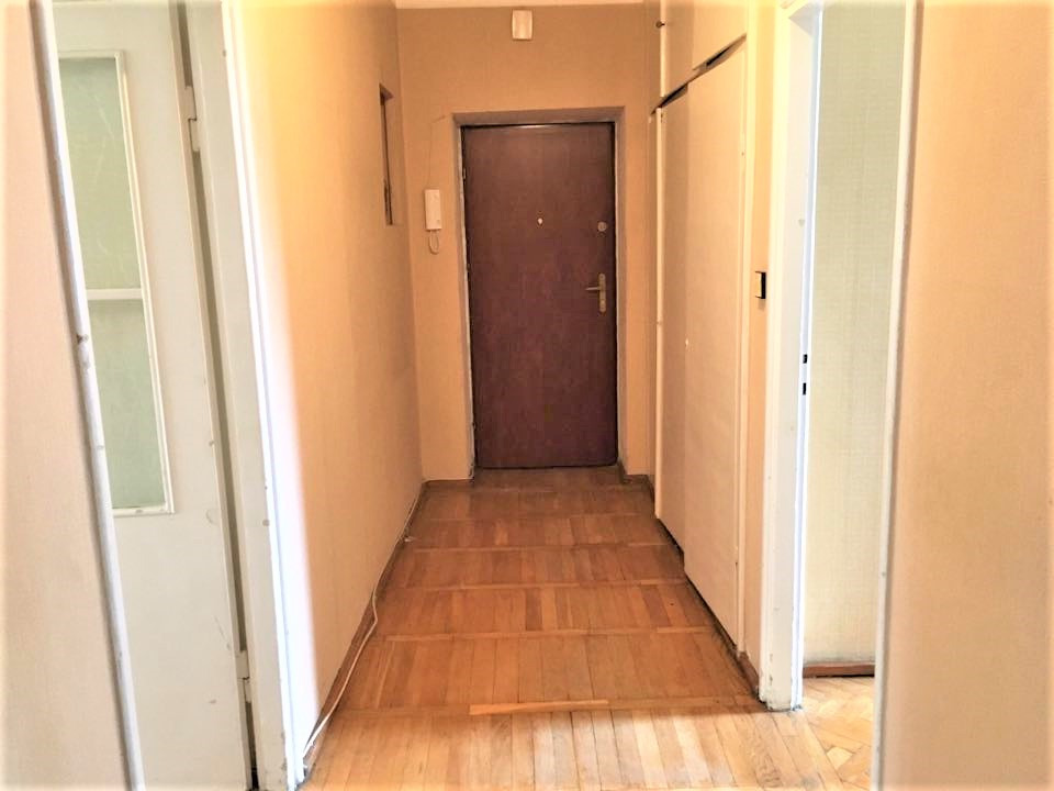 Wnętrze korytarza mieszkania z parkietem, brązowymi drzwiami wejściowymi i białymi drzwiami wewnętrznymi po obu stronach.