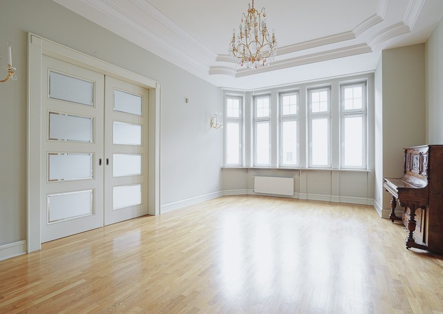 Jasne, przestronne wnętrze pokoju z parkietem, dużym oknem wykuszowym, przesuwnymi drzwiami z matowego szkła i starym pianinem.