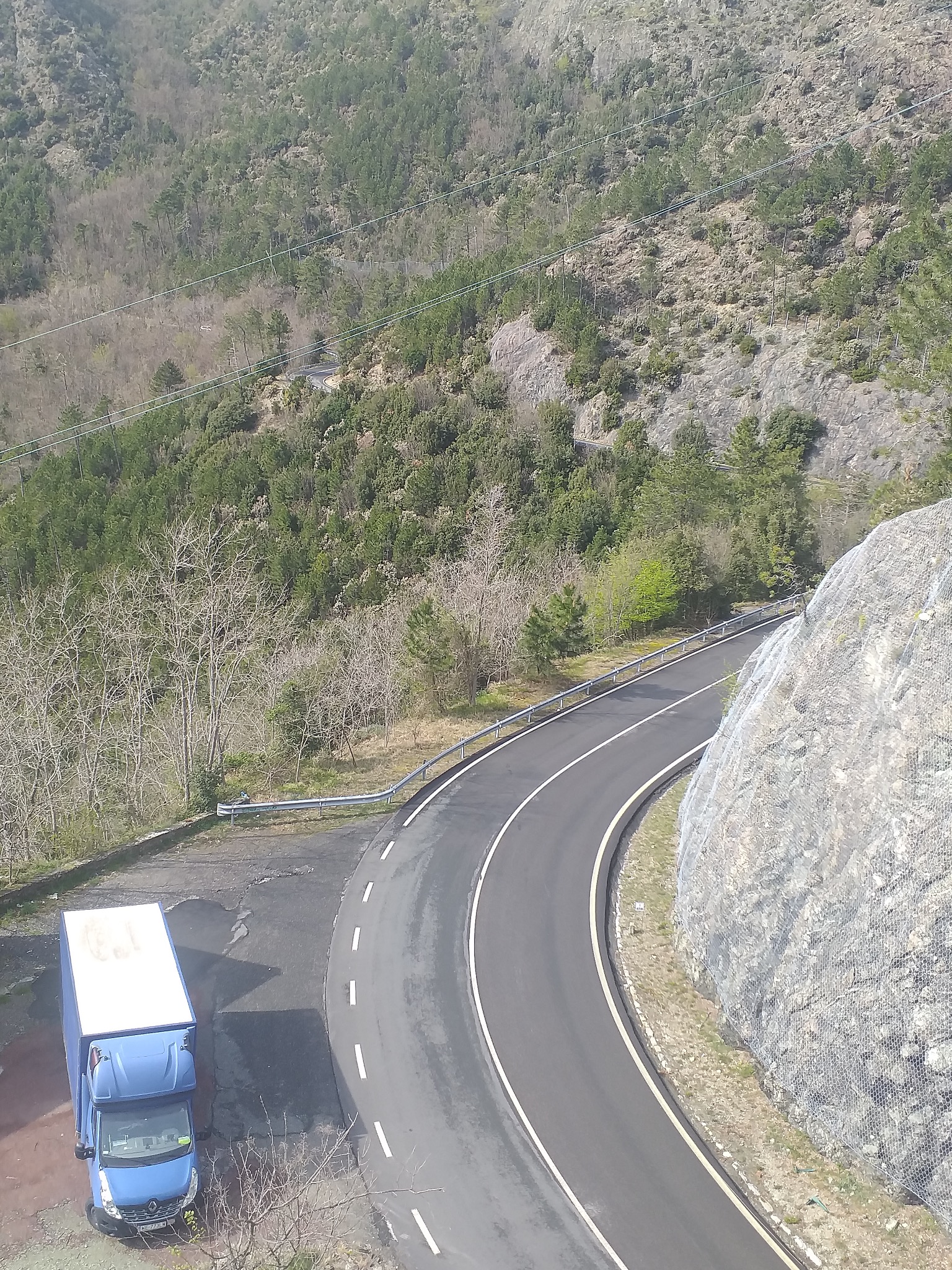 Niebieski bus z białą zabudową stoi na poboczu zakręcającej drogi górskiej z zabezpieczeniem skalnym, widok z góry.