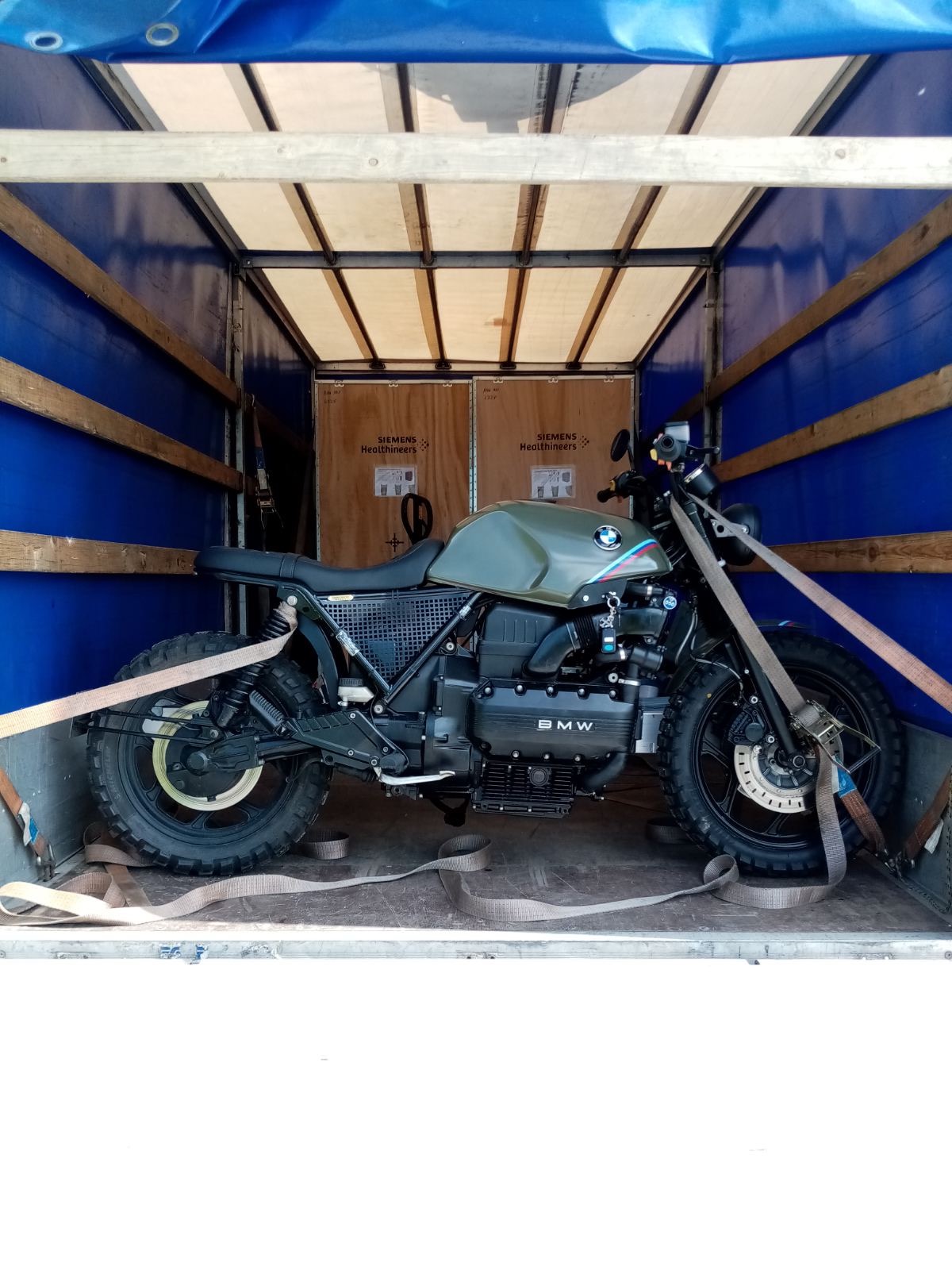 Motocykl BMW K100 w kolorze khaki zabezpieczony pasami wewnątrz skrzyni ładunkowej busa, widoczne drewniane skrzynie z napisem Siemens Healthineers w tle.