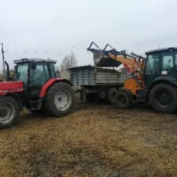 Czerwony traktor i żółto-czarna koparko-ładowarka w trakcie załadunku ziemi na przyczepę w pochmurny dzień.