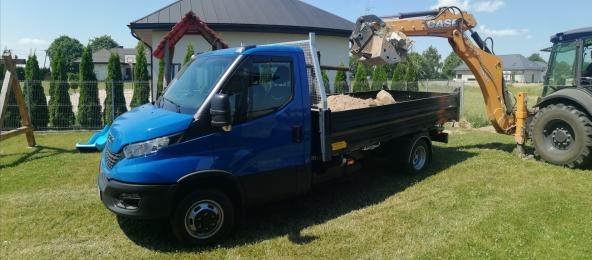 Niebieski samochód dostawczy z otwartą skrzynią ładunkową, napełniany ziemią przez koparko-ładowarkę CASE, na tle zadbanego trawnika i domu jednorodzinnego z elementami placu zabaw.